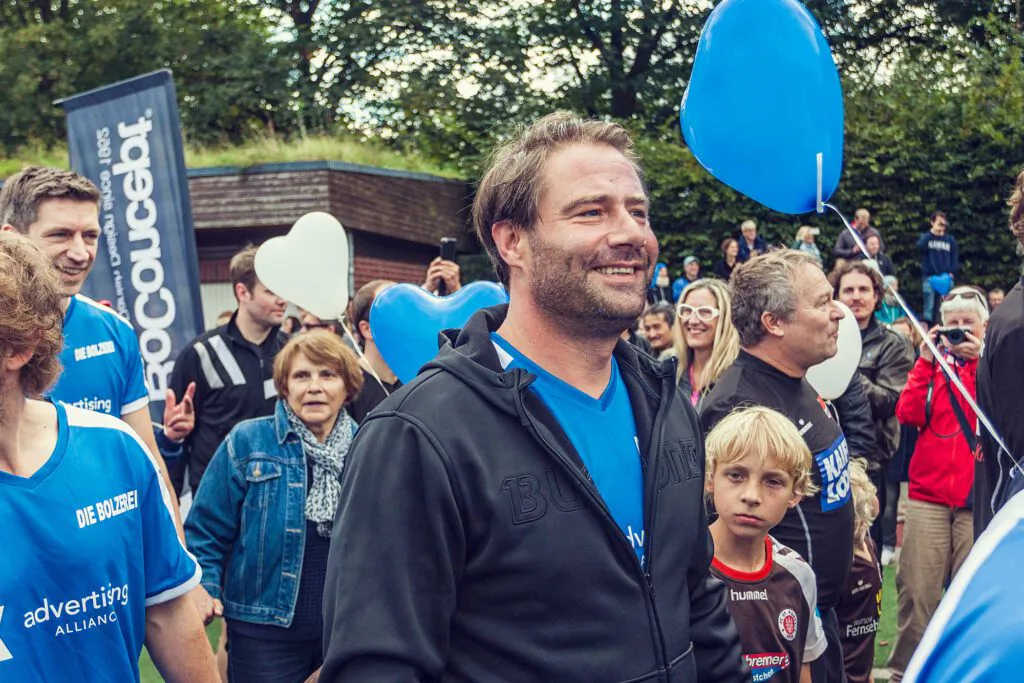 Ein lächelnder Mann in schwarzer Jacke steht mit einem Jungen in einer Menschenmenge mit blauen und weißen Herzballons.