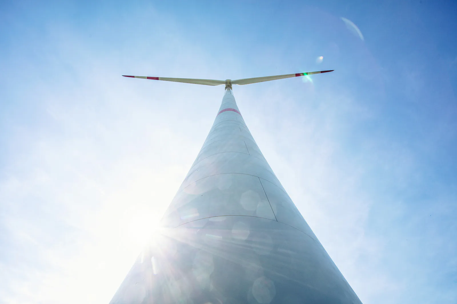 Windkraftanlage aus steiler Untersicht, mit Rotorblättern vor blauem Himmel und Sonnenstrahlen am Turm.