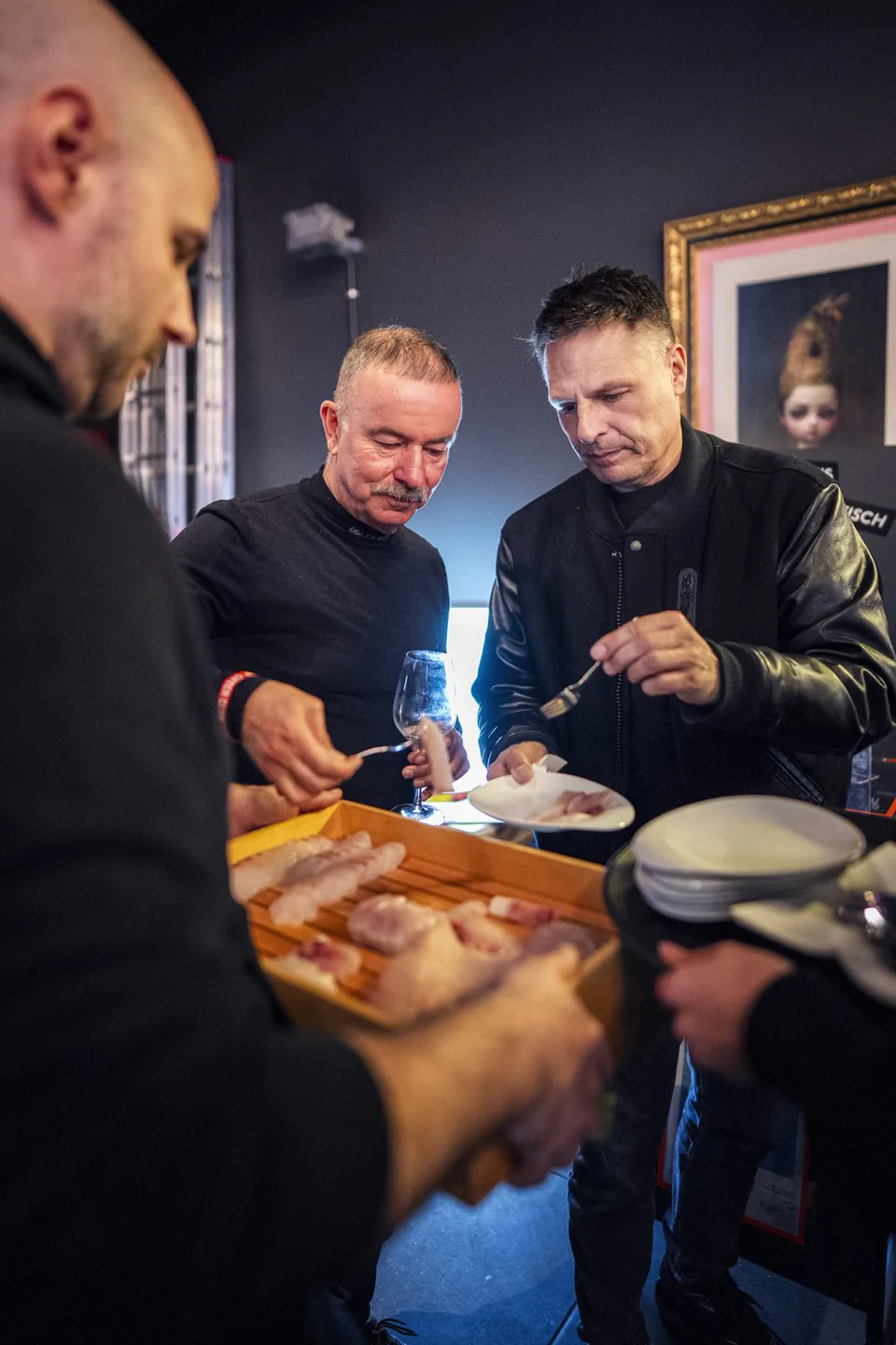 Männer verkosten Fischhäppchen von einem Holztablett in einem dunkel gestrichenen Innenraum mit gerahmten Bildern.