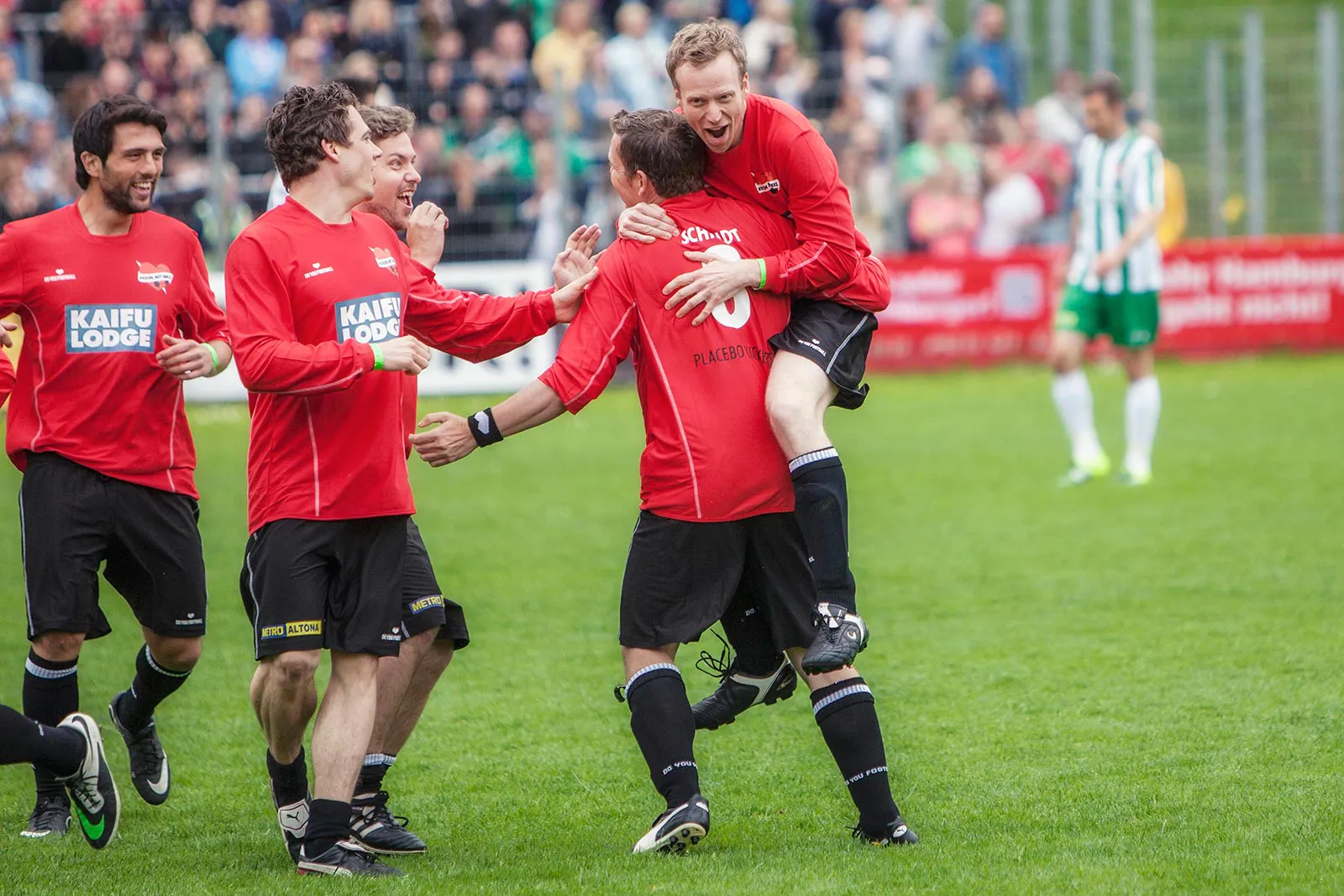 Fußballspieler in roten Trikots jubeln auf dem Rasen; einer springt einem Mitspieler vor Zuschauern in die Arme.