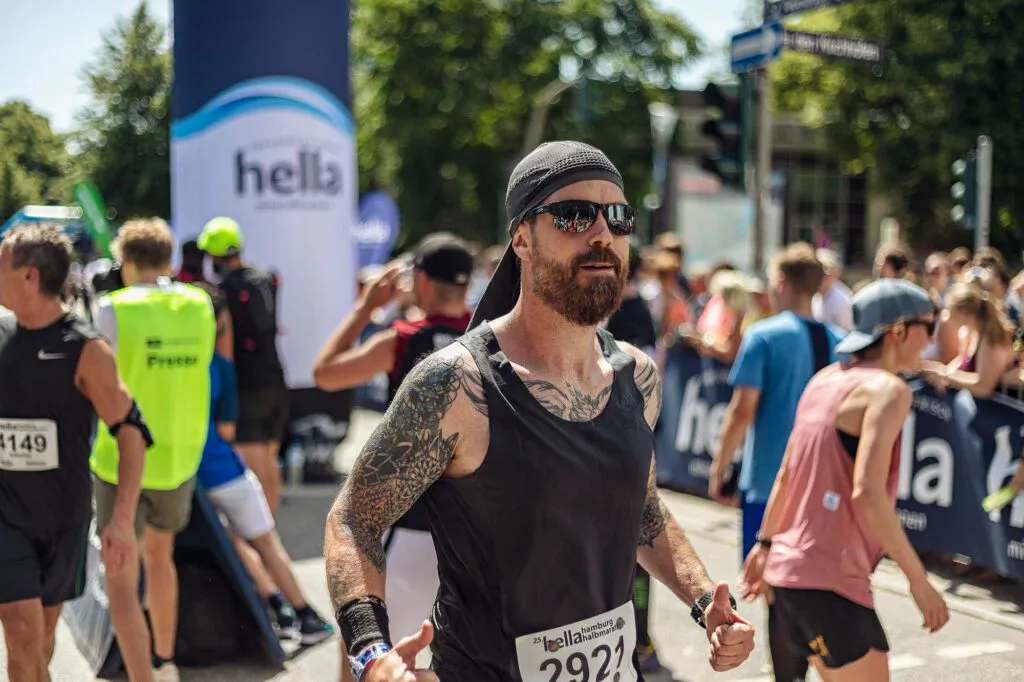 Tätowierter Läufer mit Sonnenbrille und schwarzem Tanktop zwischen anderen Teilnehmenden auf sonniger Straße.