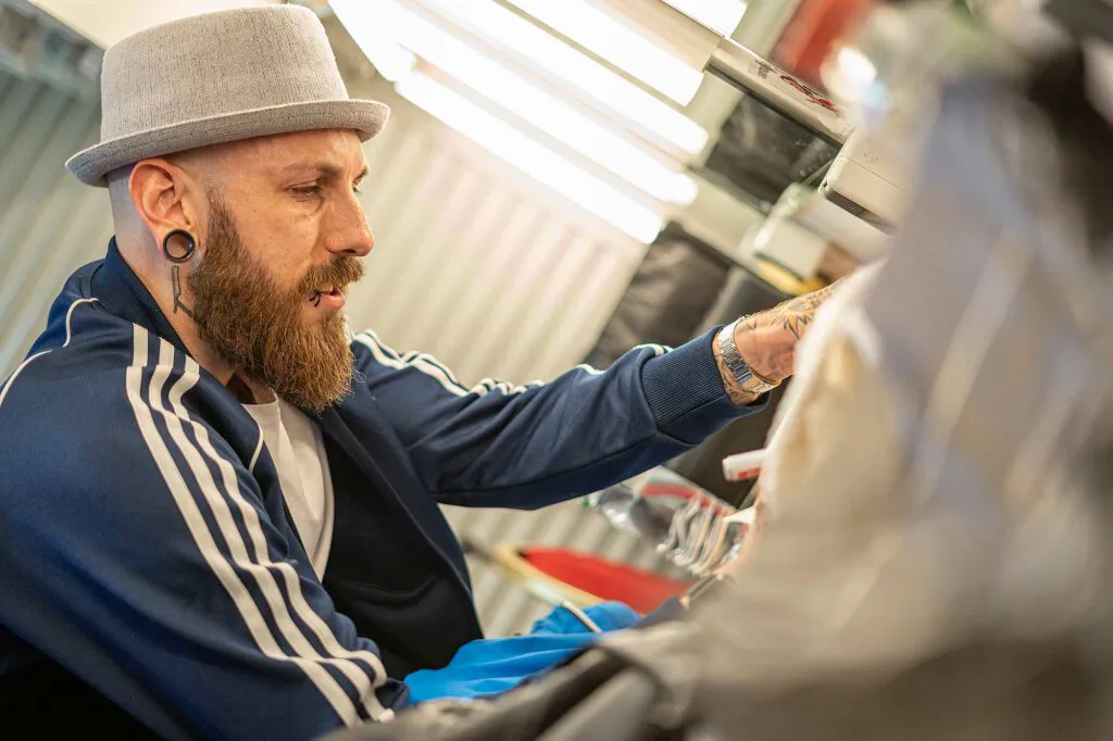 Dennis Grundt mit grauem Hut, Bart und blauer Trainingsjacke in einem hell beleuchteten Raum.