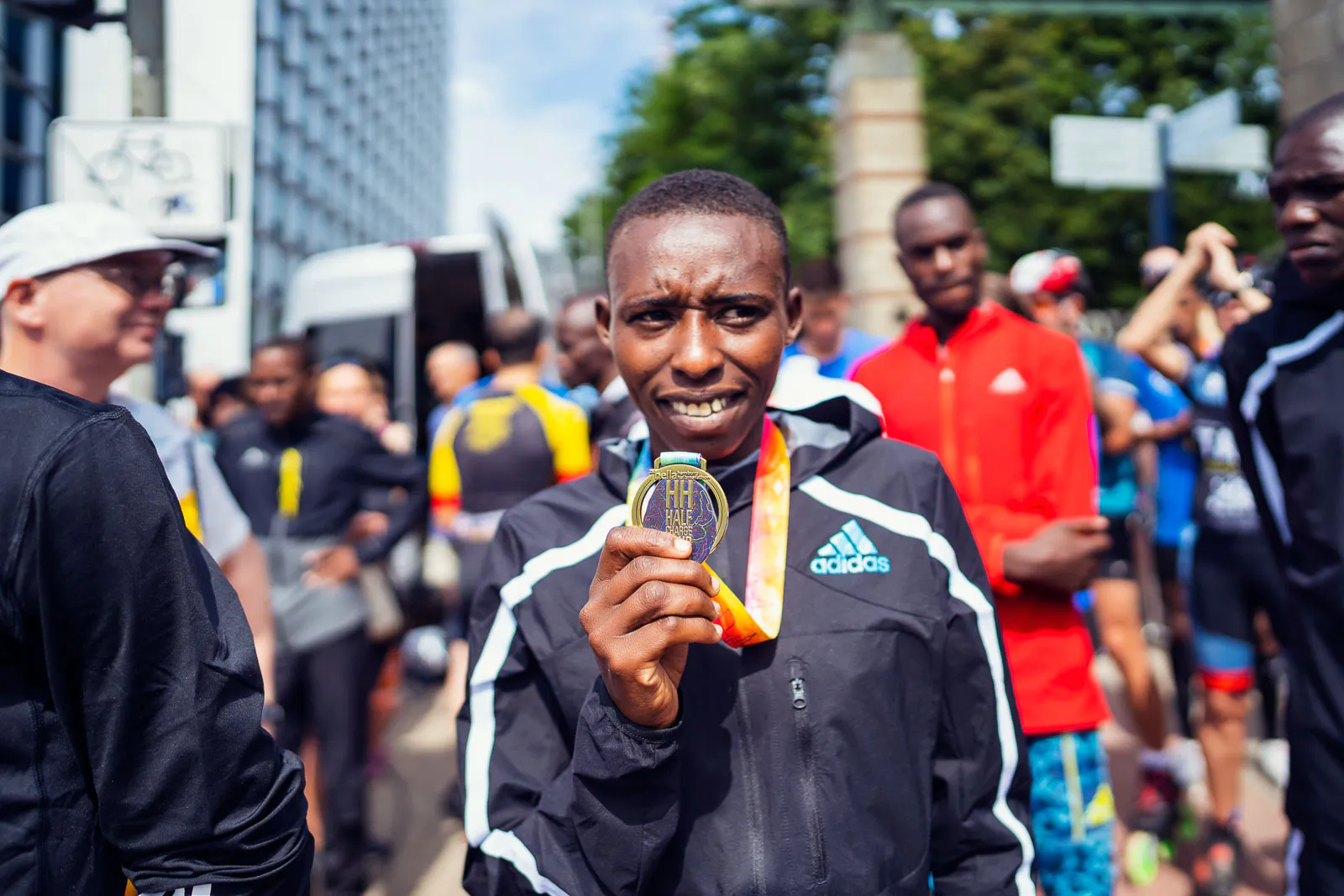 Läufer in schwarzer Sportjacke hält seine Medaille vor die Kamera, umgeben von Menschen auf sonniger Straße.