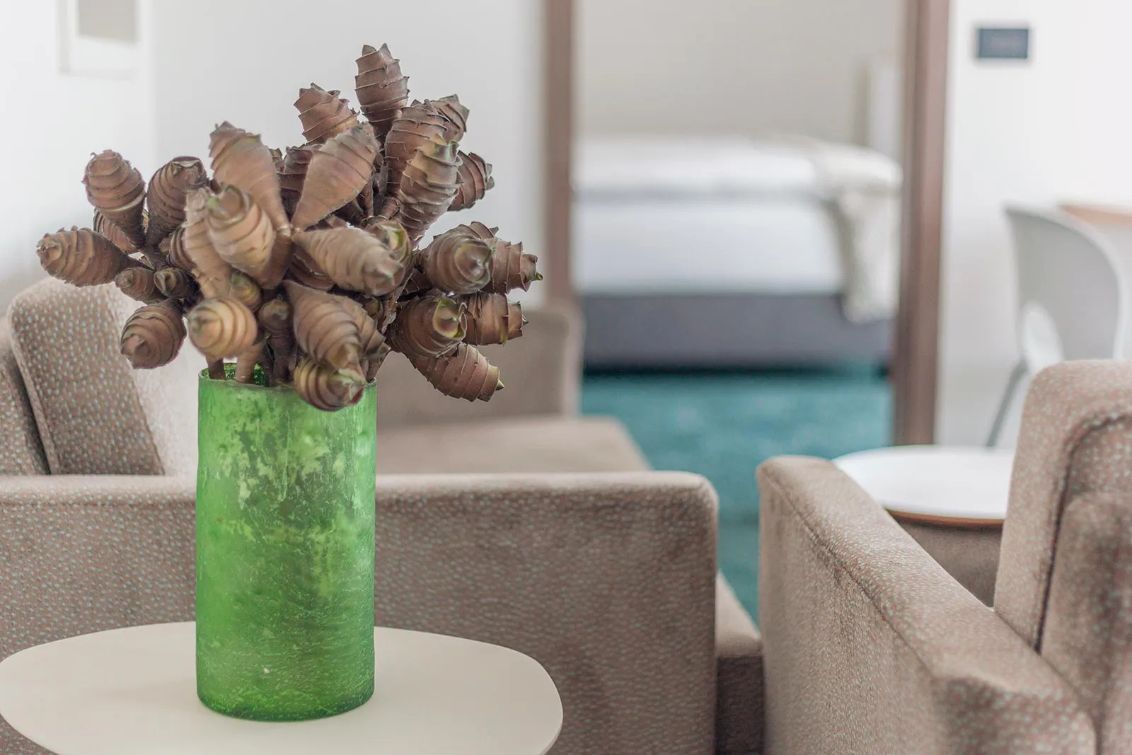 Grüne Vase mit braunem Pflanzenschmuck auf einem Tisch zwischen Sesseln, dahinter ein Hotelbett im Tageslicht.