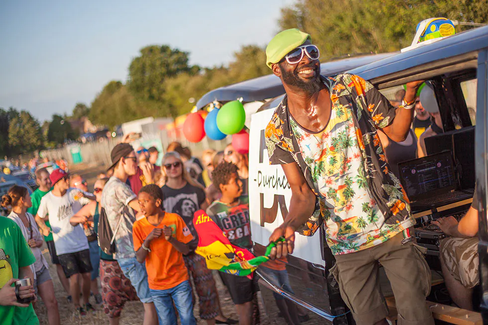 Mann mit grüner Mütze, Sonnenbrille und Blumenhemd neben einem Fahrzeug in sonniger Festivalmenge.