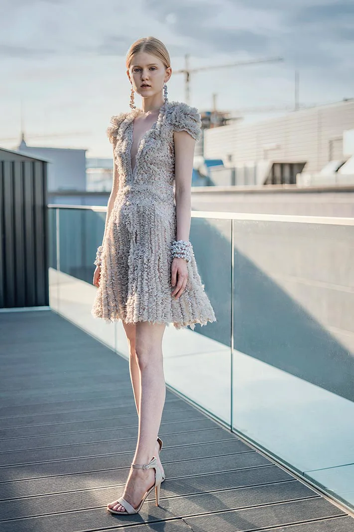 Frau in strukturiertem, hellem Minikleid und High Heels posiert im Sonnenlicht auf einer Dachterrasse mit Glasgeländer.
