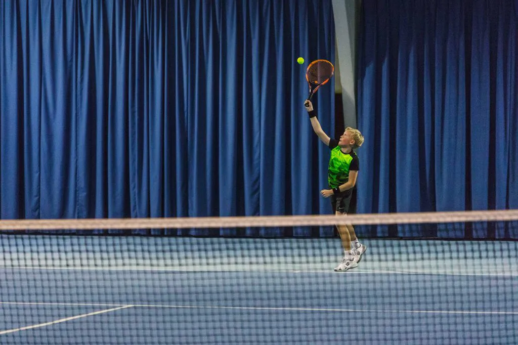 Junger Tennisspieler im grünen Shirt beim Aufschlag in einer Tennishalle mit blauen Vorhängen.