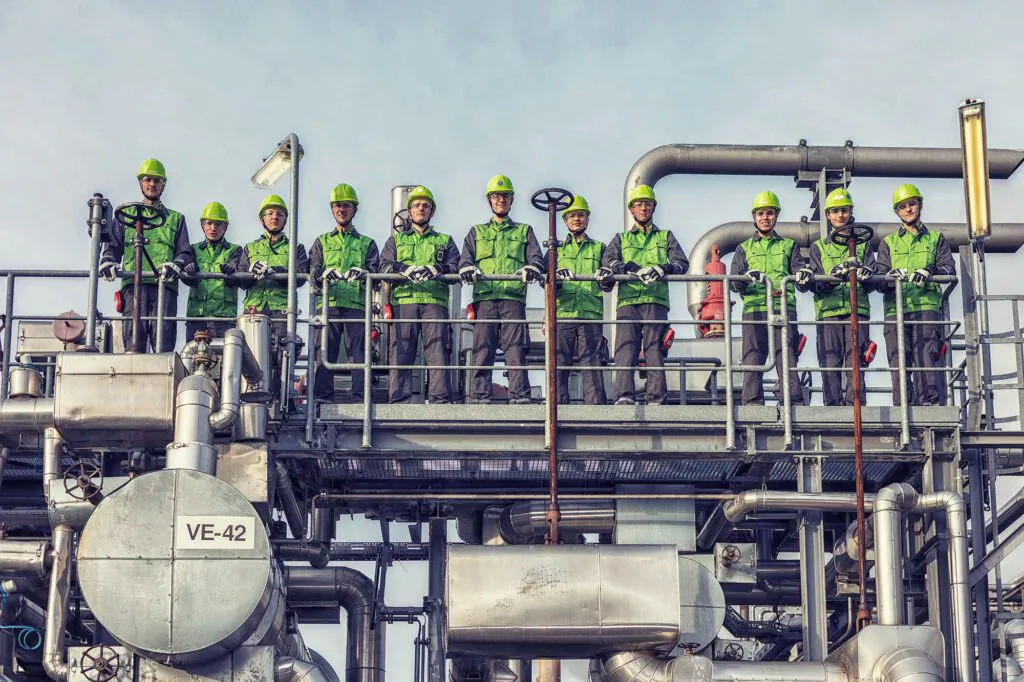 Gruppe von Arbeitern mit grünen Schutzhelmen steht auf einer Plattform zwischen Rohrleitungen der Raffinerie Heide.