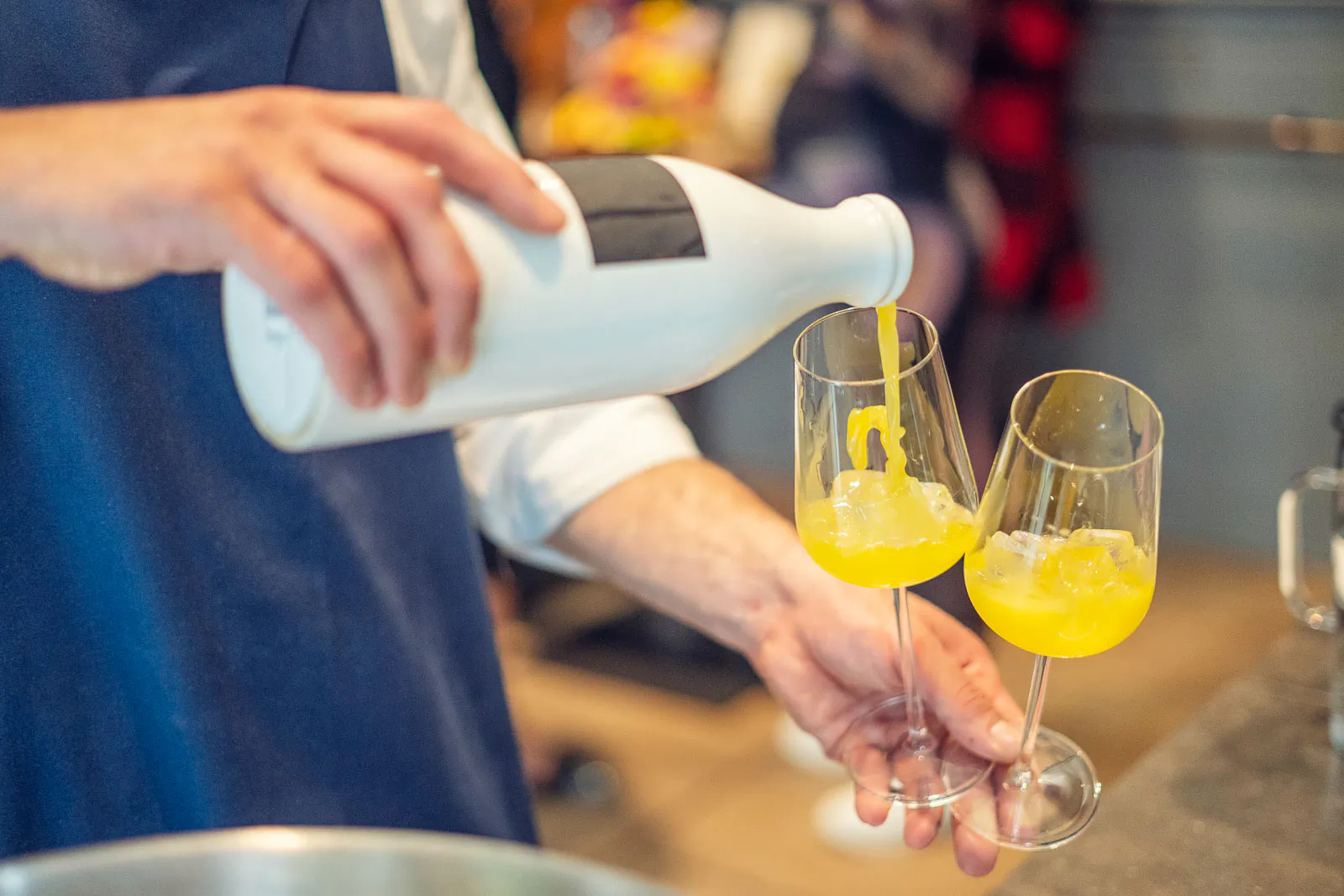 Person in blauer Schürze gießt ein gelbes Getränk aus einer weißen Flasche in zwei Weingläser mit Eis.
