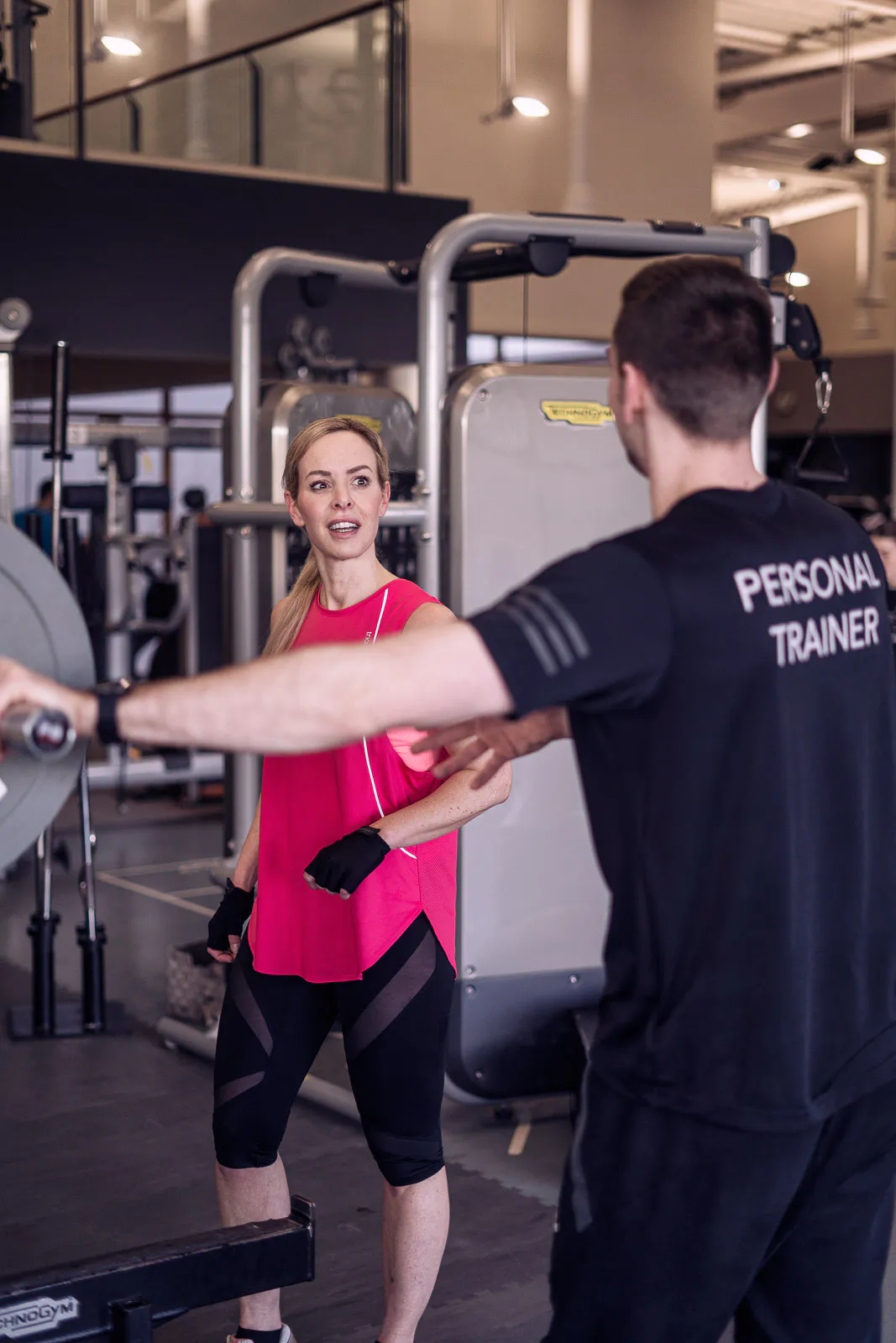 Isabel Edvardsson im pinken Sporttop mit Personal Trainer zwischen Trainingsgeräten im Fitnessstudio.