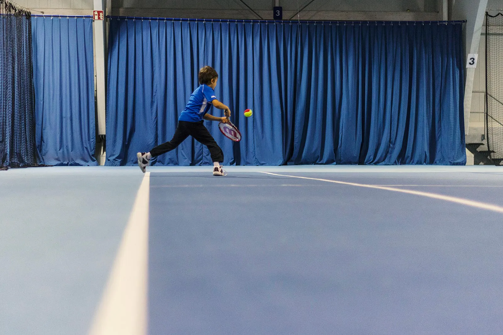 Kind im blauen Shirt schlägt einen Tennisball in einer Tennishalle vor blauem Vorhang.