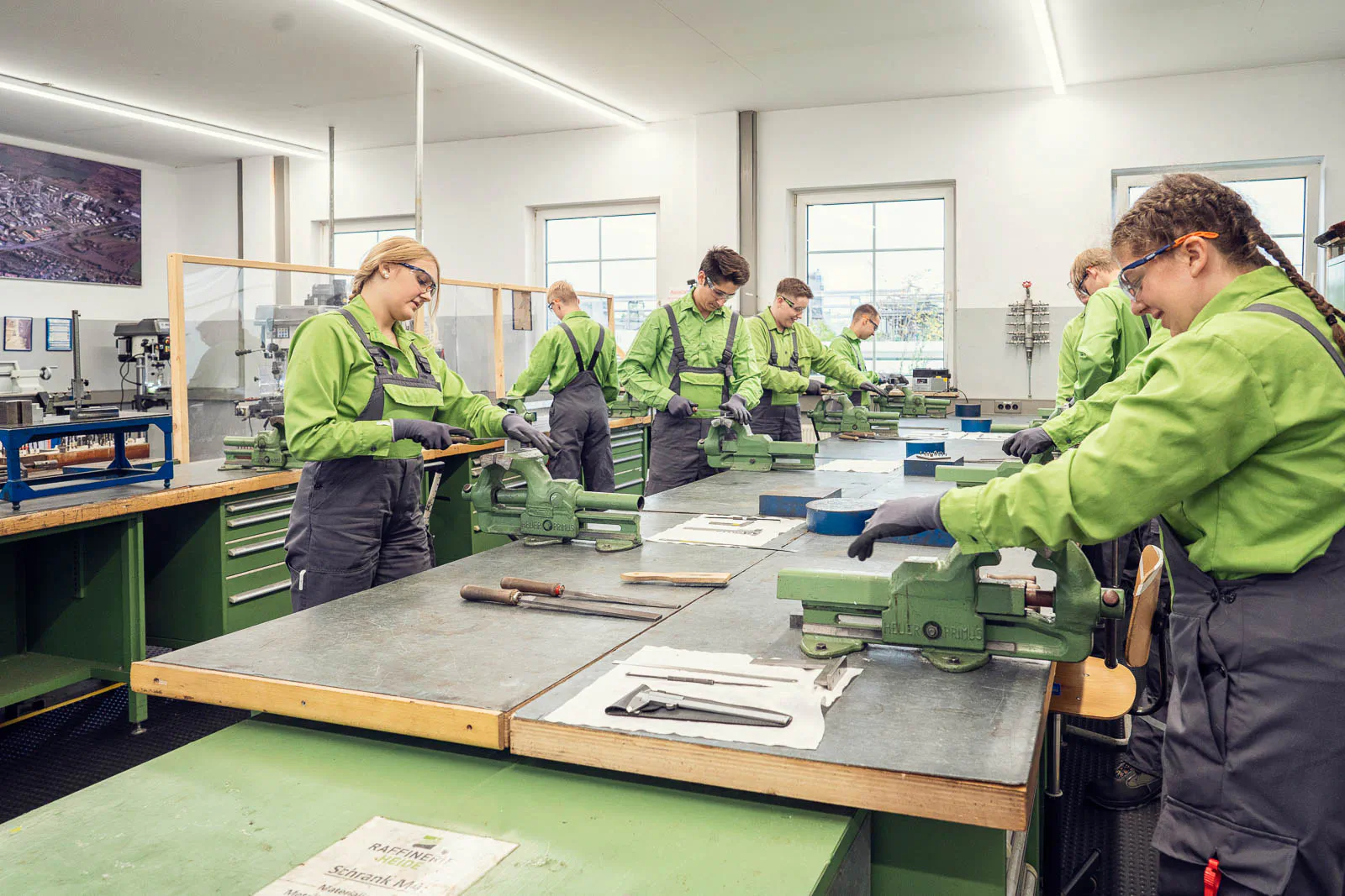Azubis in grüner Arbeitskleidung bearbeiten Werkstücke an Schraubstöcken in einer hellen Werkstatt der Raffinerie Heide.