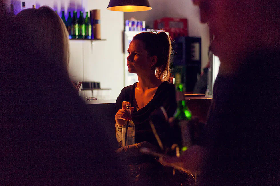 Eine Frau hält eine Flasche an einer Bar, umgeben von Gästen im warmen Licht einer Hängelampe.