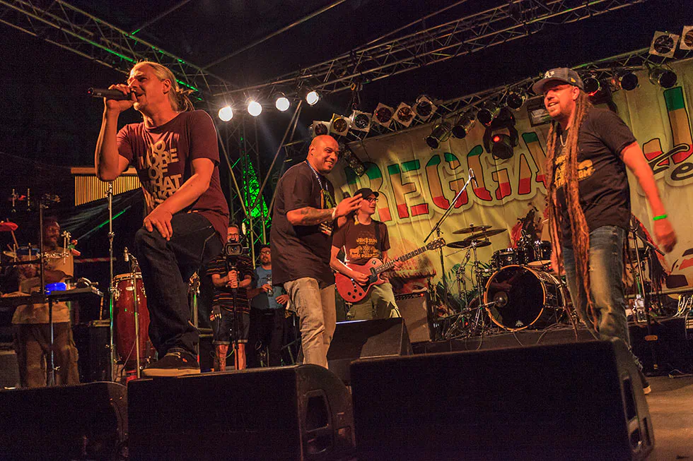 Sänger mit Mikrofonen und Gitarrist auf der beleuchteten Reggae-Jam-Bühne, rechts ein Musiker mit langen Dreadlocks.
