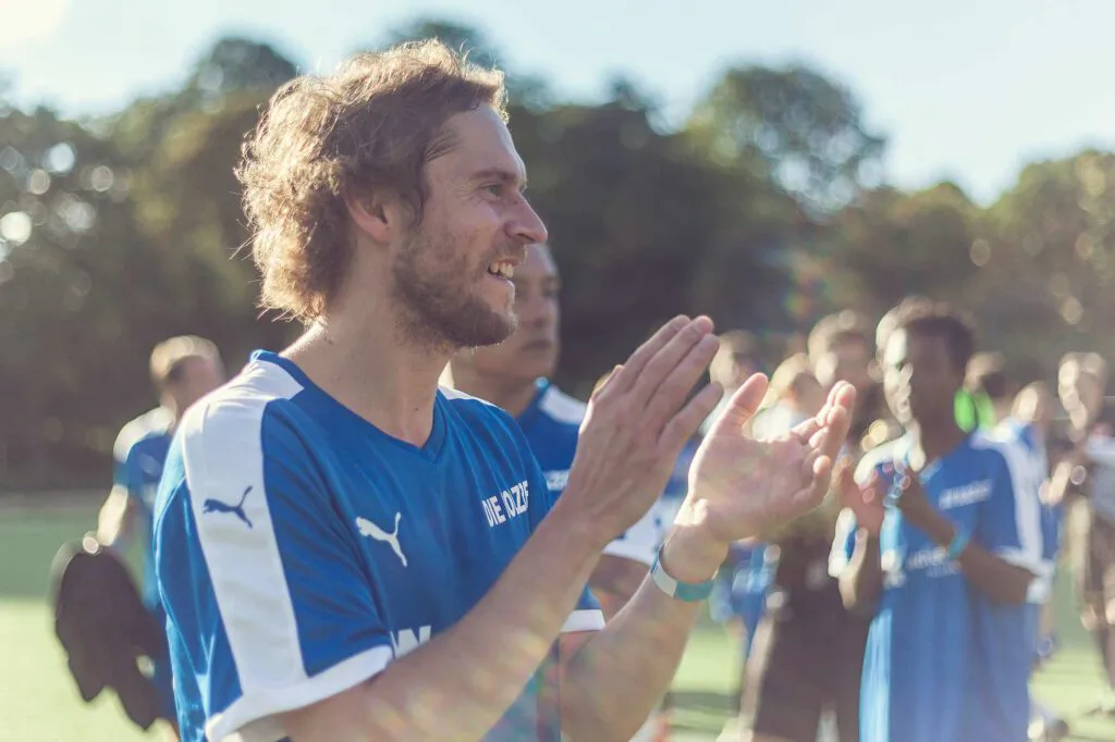 Mann im blauen Fußballtrikot klatscht auf einem sonnigen Sportplatz, weitere Spieler stehen im Hintergrund.