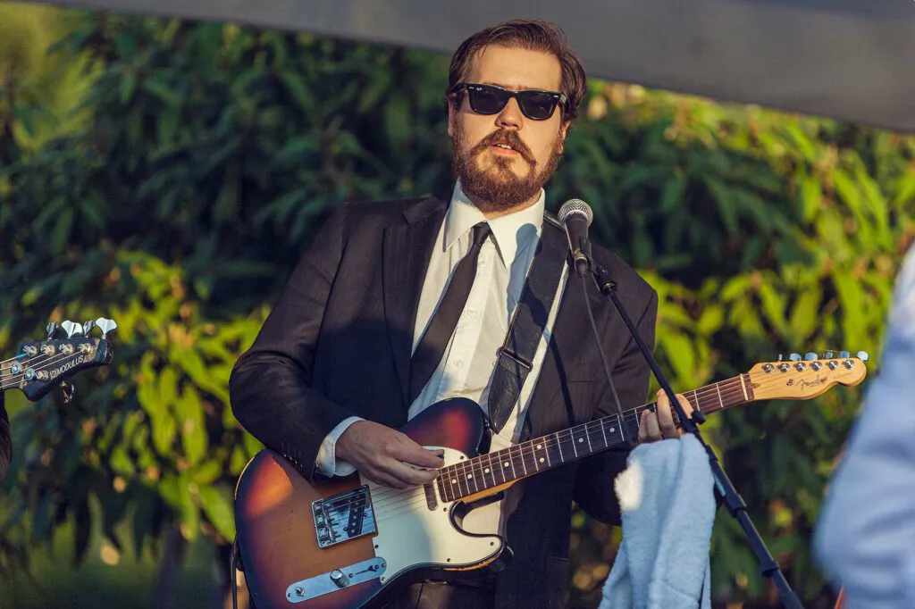 Gitarrist mit Sonnenbrille und Anzug spielt E-Gitarre am Mikrofon vor grüner Pflanzenwand im warmen Licht.