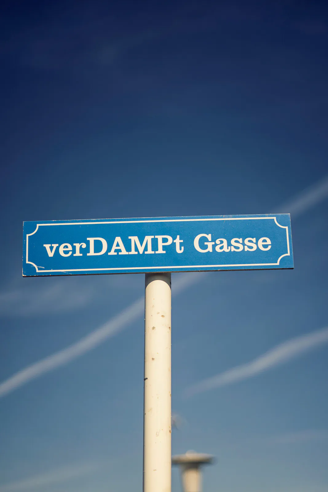Blaues Straßenschild mit der Aufschrift verDAMPt Gasse an einem hellen Pfosten vor blauem Himmel mit Kondensstreifen.