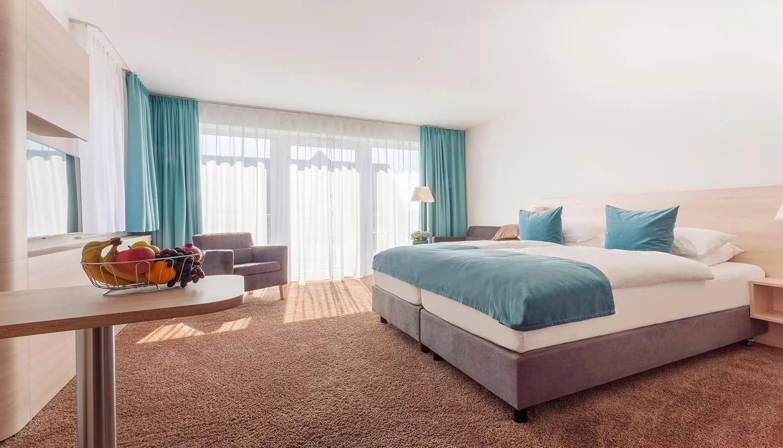 Helles Hotelzimmer mit Doppelbett, türkisfarbenen Kissen und Vorhängen, Sessel und Obstschale auf einem Tisch.