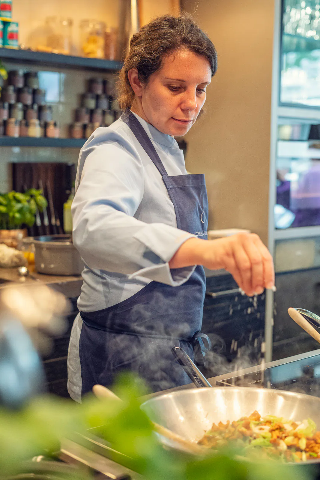 Köchin in blauer Schürze würzt eine dampfende Pfanne mit Gemüse in einer warm beleuchteten Restaurantküche.