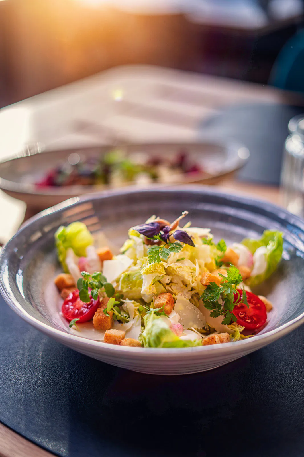 Salat mit Tomaten, Croûtons, Käsehobeln und Kräutern in einer Schale auf einem Restauranttisch im warmen Sonnenlicht.