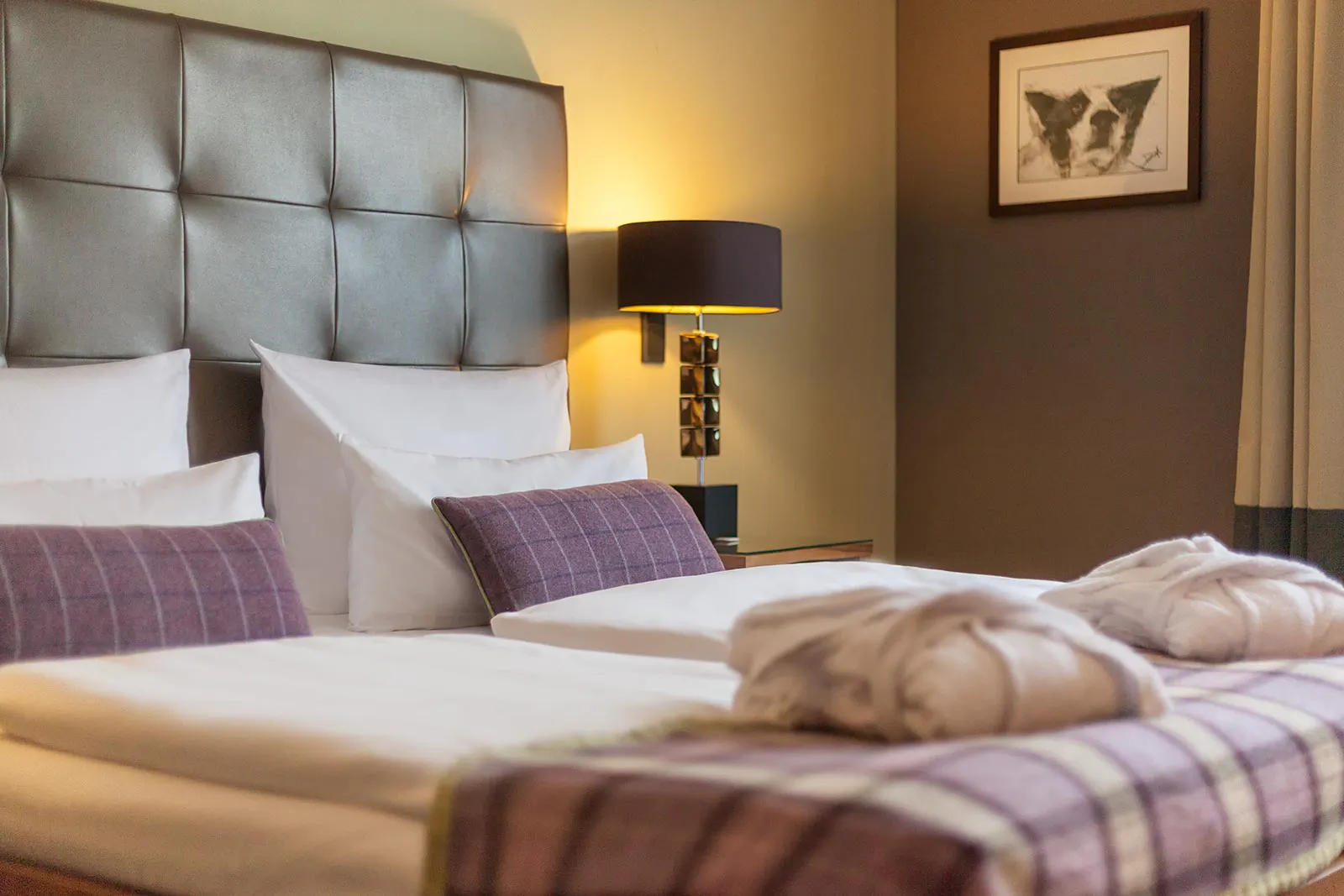 Hotelbett mit weißen Kissen, violetten Akzenten und zwei Bademänteln vor gepolstertem Kopfteil im warmen Lampenlicht.