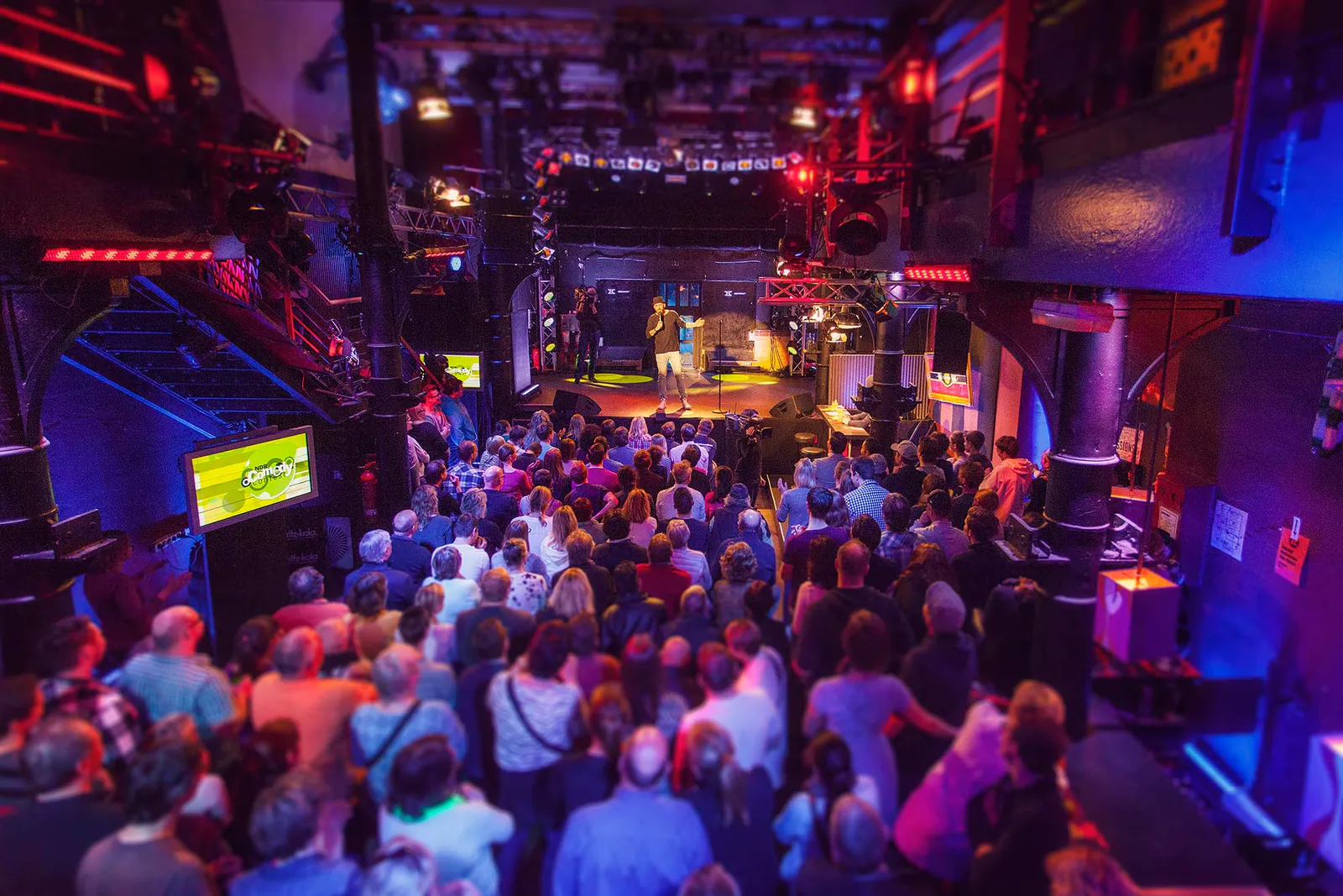 Publikum vor einer kleinen Bühne beim NDR Comedy Contest, getaucht in rotes und blaues Licht.