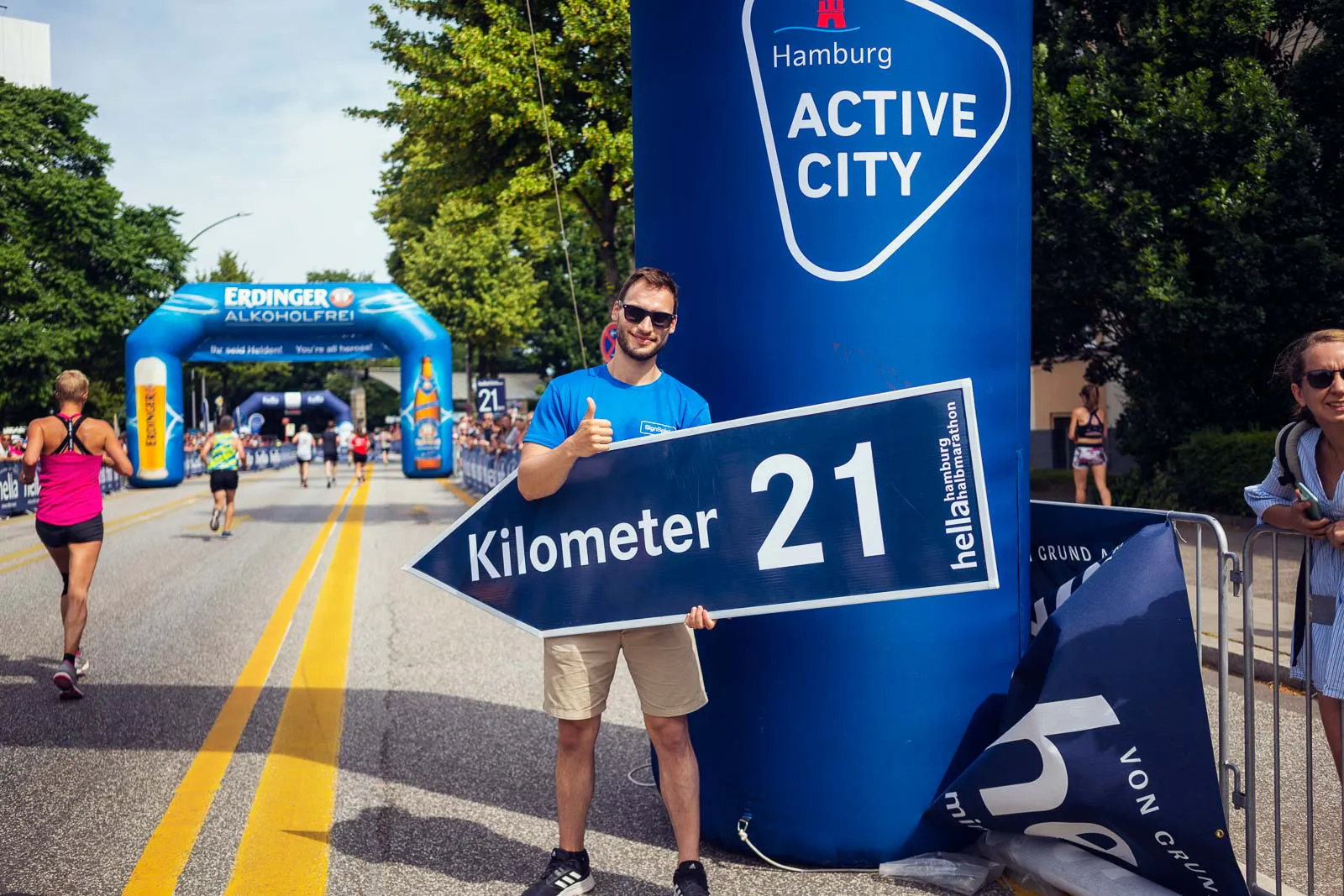 Mann mit Sonnenbrille hält ein Kilometer-21-Schild am sonnigen Straßenrand, während Läufer vorbeiziehen.
