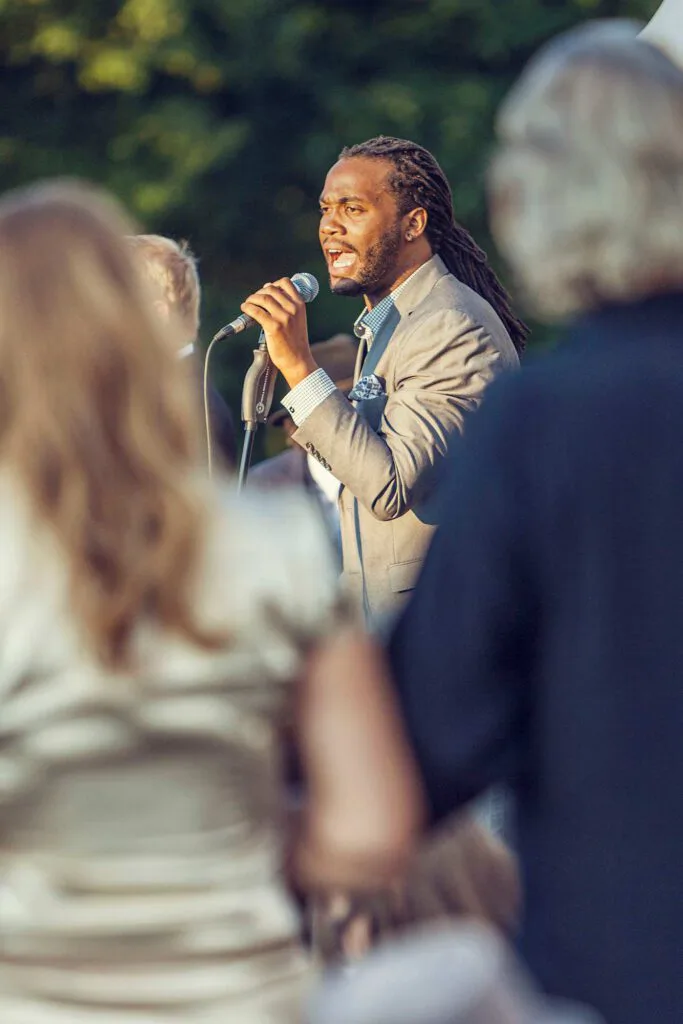 Sänger mit Dreadlocks und grauem Sakko singt im warmen Sonnenlicht ins Mikrofon, vor ihm Gäste im Freien.