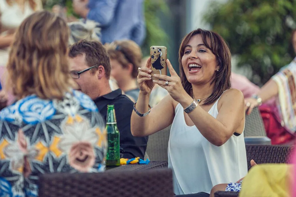 Lachende Frau im weißen Oberteil hält ein Smartphone hoch, umgeben von Gästen an Tischen auf einer Terrasse.