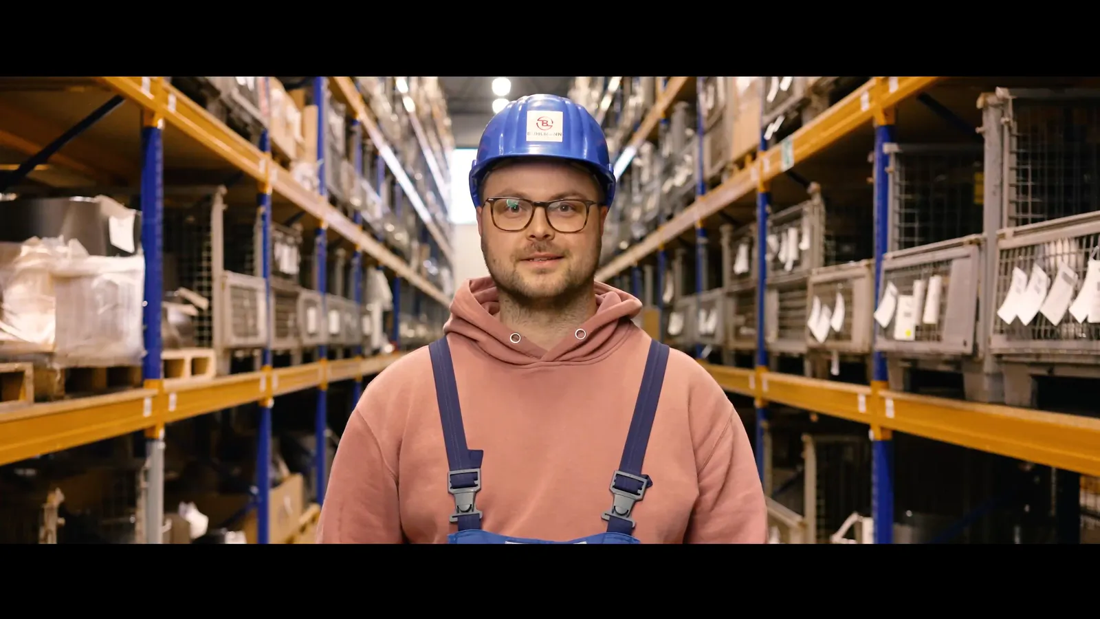 Mann mit blauem Schutzhelm, Brille und rosa Hoodie steht zwischen hohen Regalen in einer beleuchteten Lagerhalle.