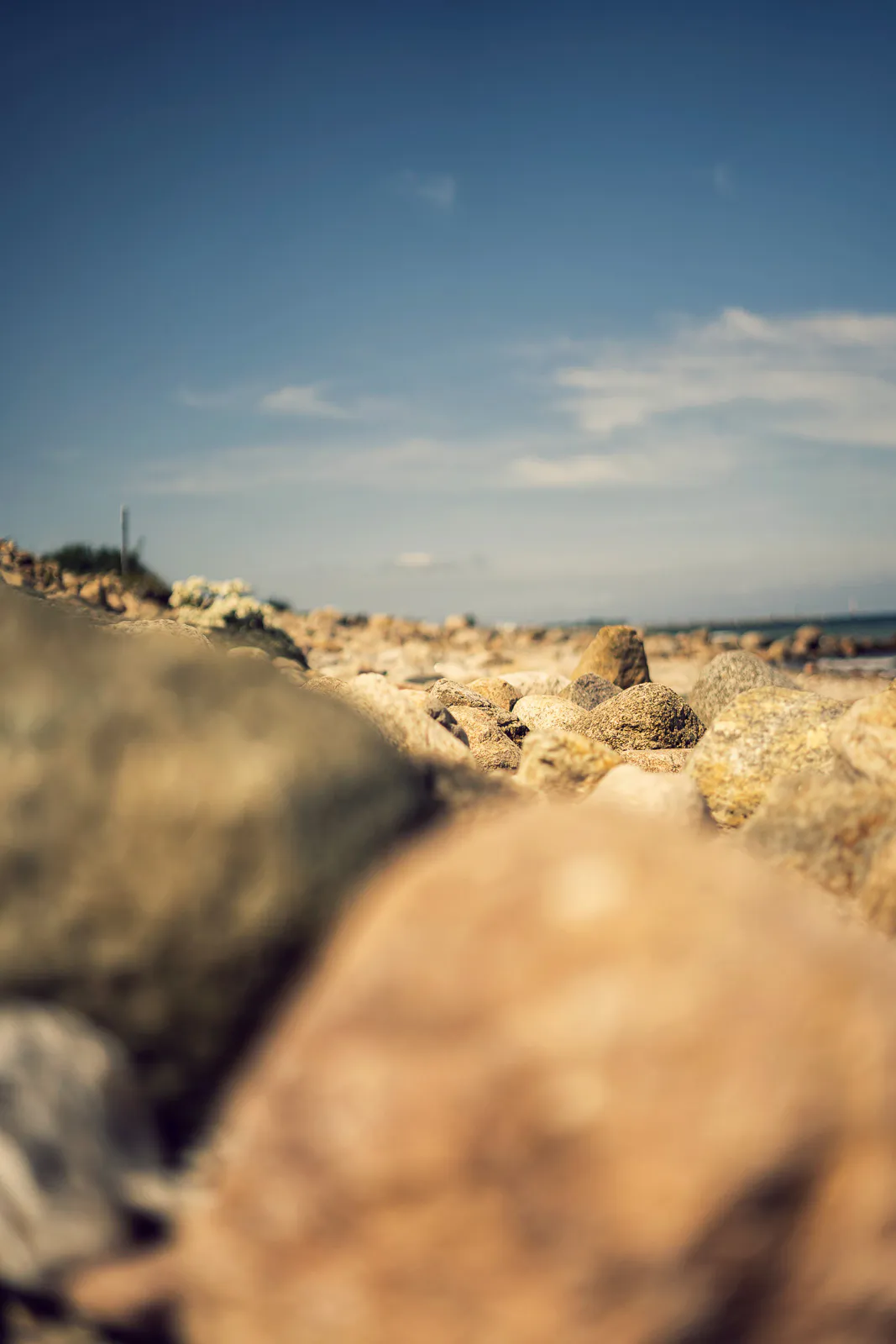 Sonniger Steinstrand aus bodennaher Perspektive, mit unscharfen Felsen im Vordergrund und blauem Himmel.