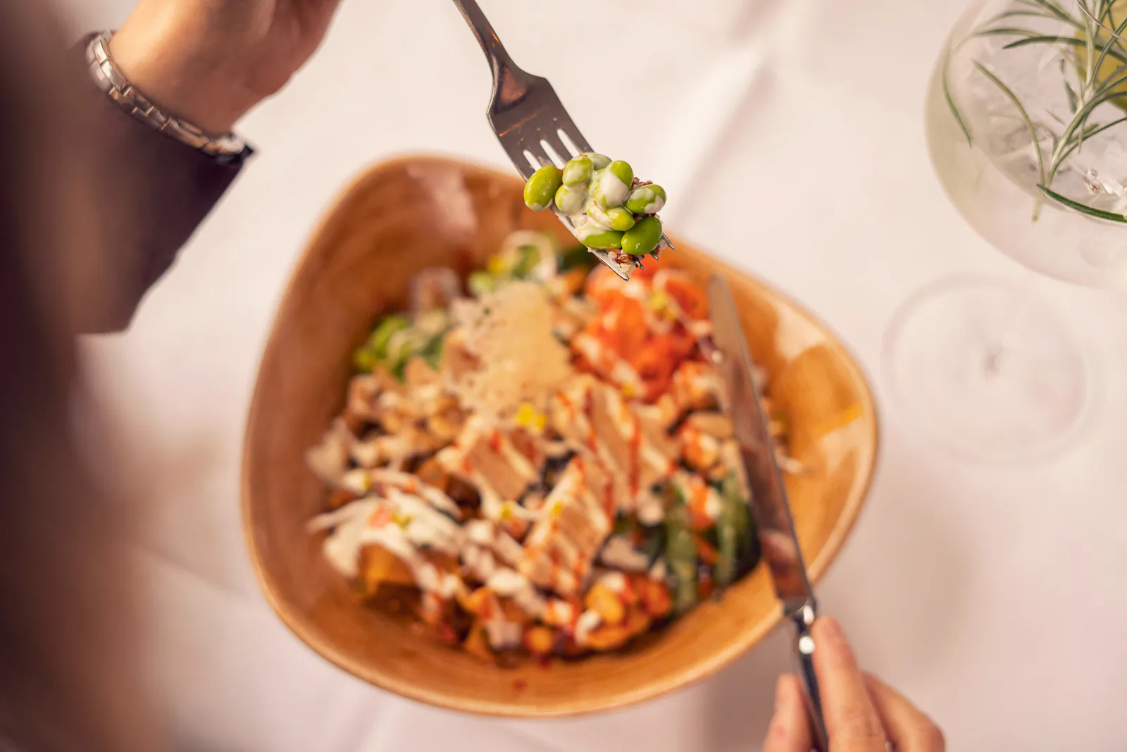 Person hält eine Gabel mit grünen Bohnen über eine bunte Bowl; daneben ein Drink mit Rosmarin auf weißem Tisch.