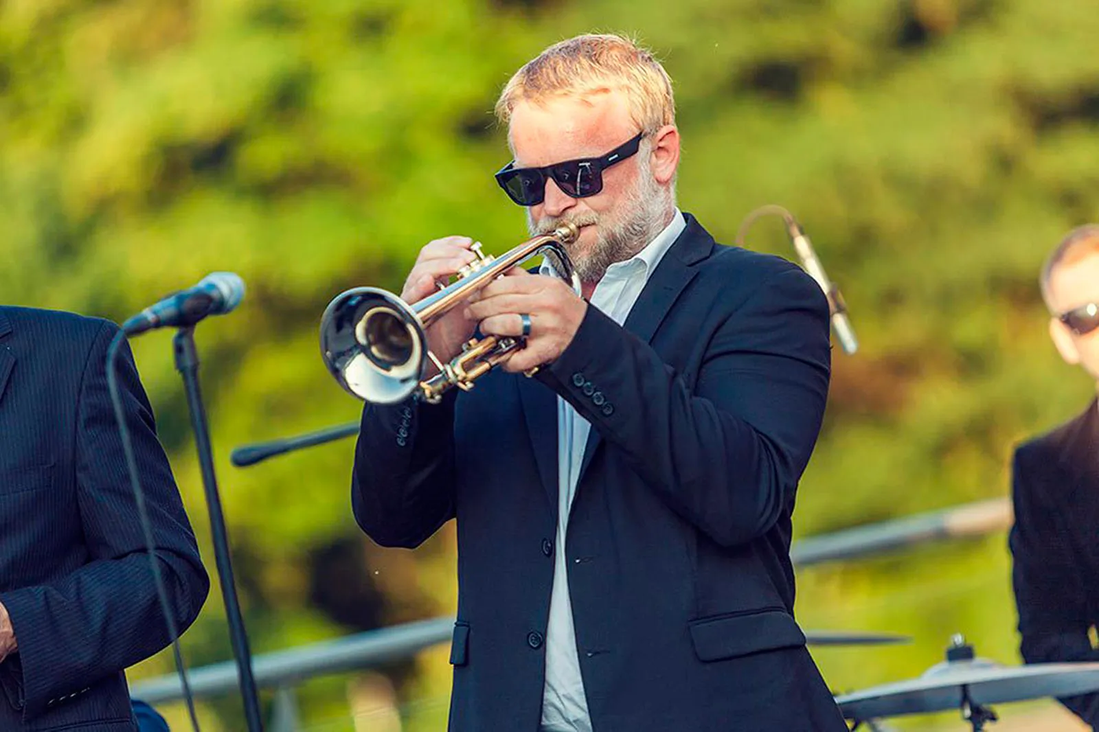 Mann mit Sonnenbrille und dunklem Sakko spielt Trompete auf einer Außenbühne vor sonnenbeschienenen Bäumen.
