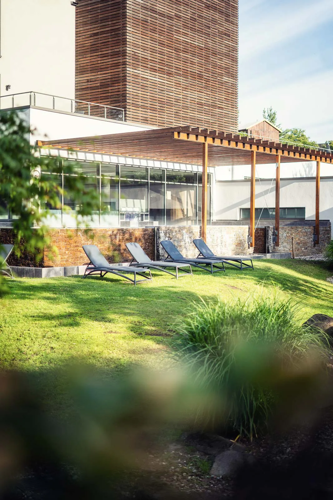 Vier Sonnenliegen auf einer sonnigen Rasenfläche vor einem Saunagebäude mit Glasfront und Holzpergola.