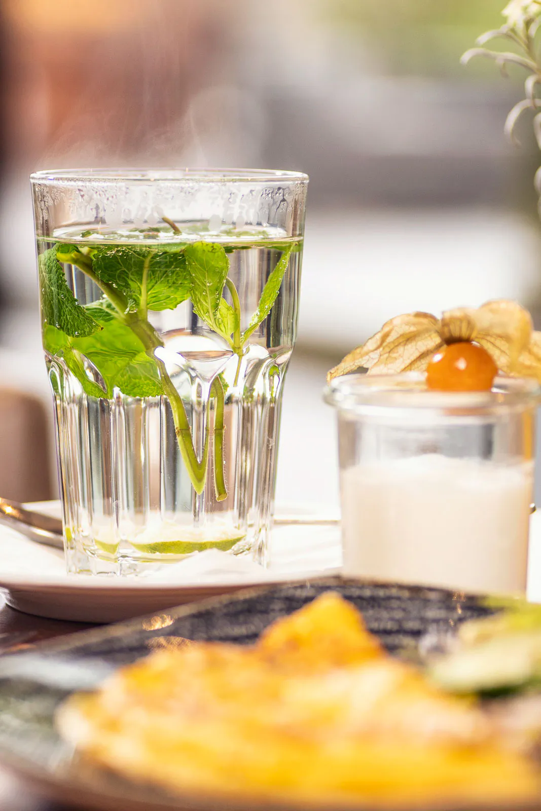 Dampfender Minztee im Glas neben einem Dessert mit Physalis und unscharfem Speiseteller im warmen Licht.