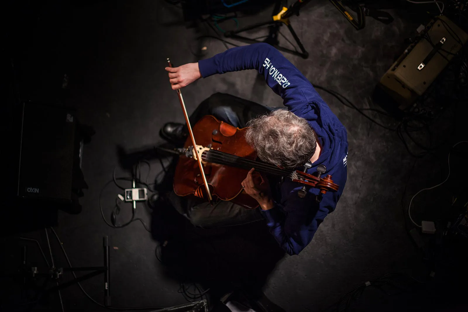 Musiker im blauen Pullover spielt Geige auf einer dunklen Bühne, von oben im Scheinwerferlicht aufgenommen.