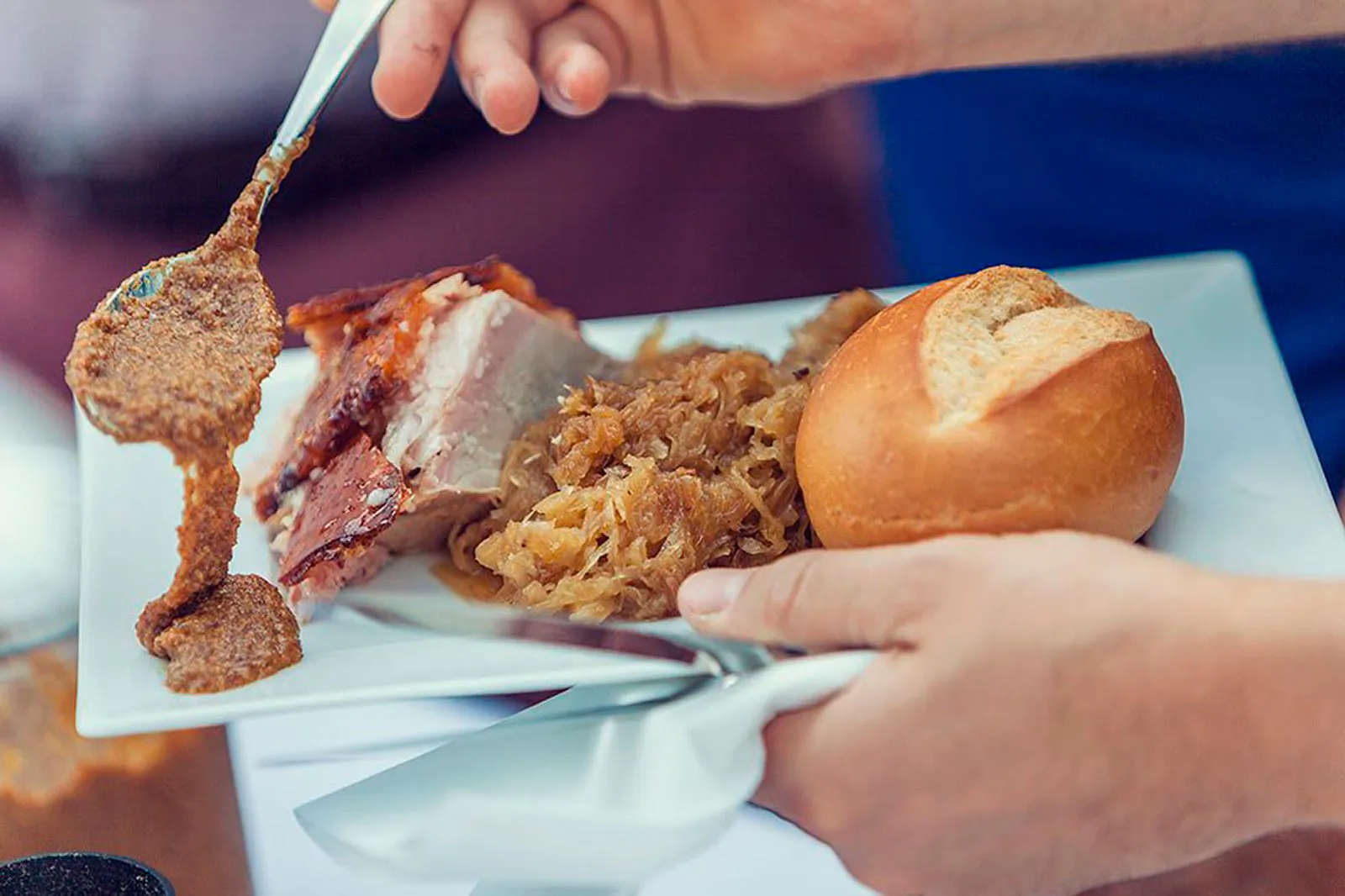 Hände halten einen Teller mit Braten, Sauerkraut und Brötchen, ein Löffel gibt Soße dazu.