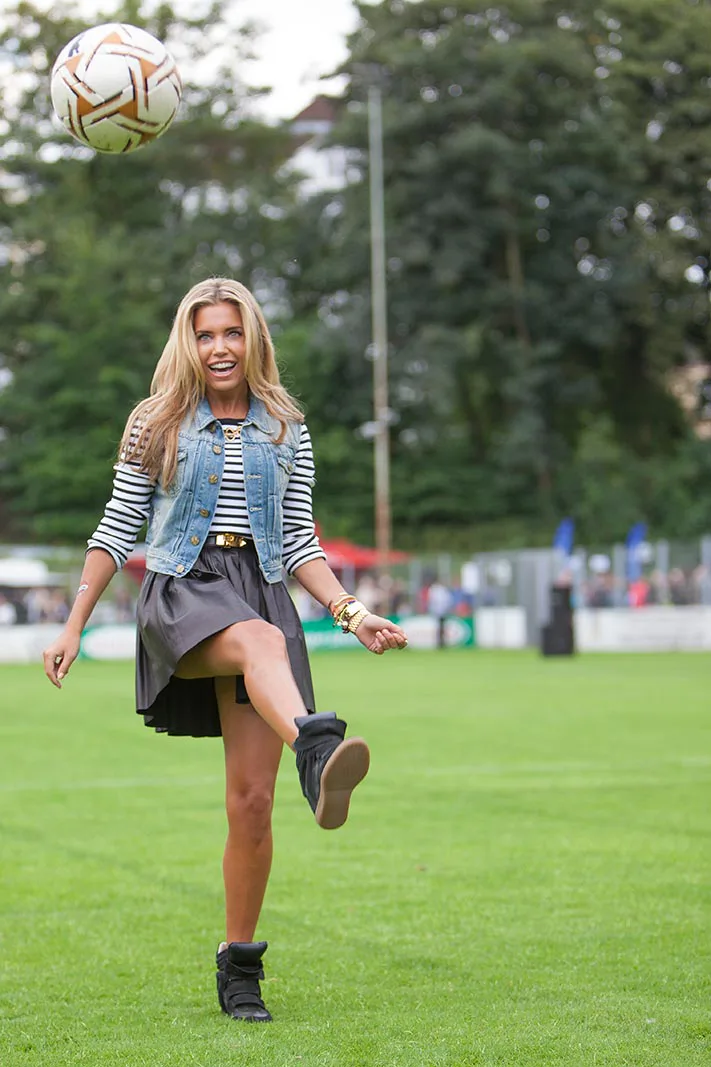Eine lachende Frau in Jeansweste und Rock kickt einen Fußball auf dem Rasen bei Kicken mit Herz.