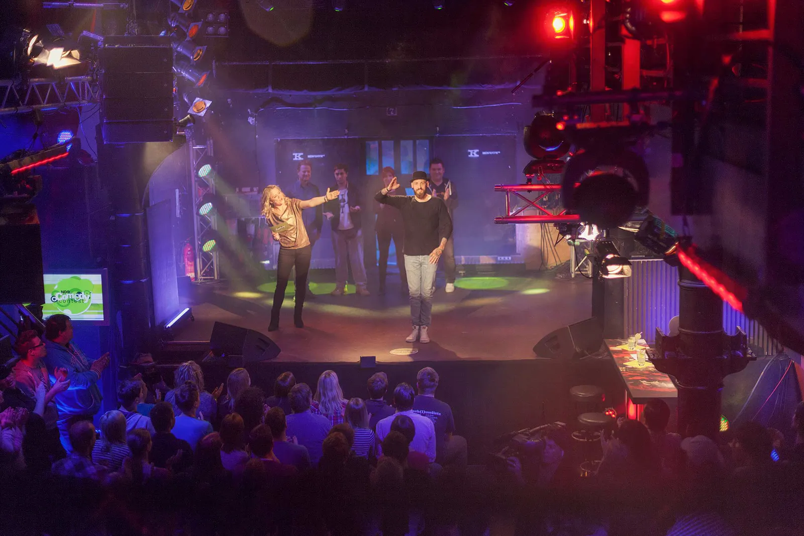 Dennis Grundt winkt auf der bunt beleuchteten Bühne des NDR Comedy Contests vor Publikum, weitere Personen hinter ihm.