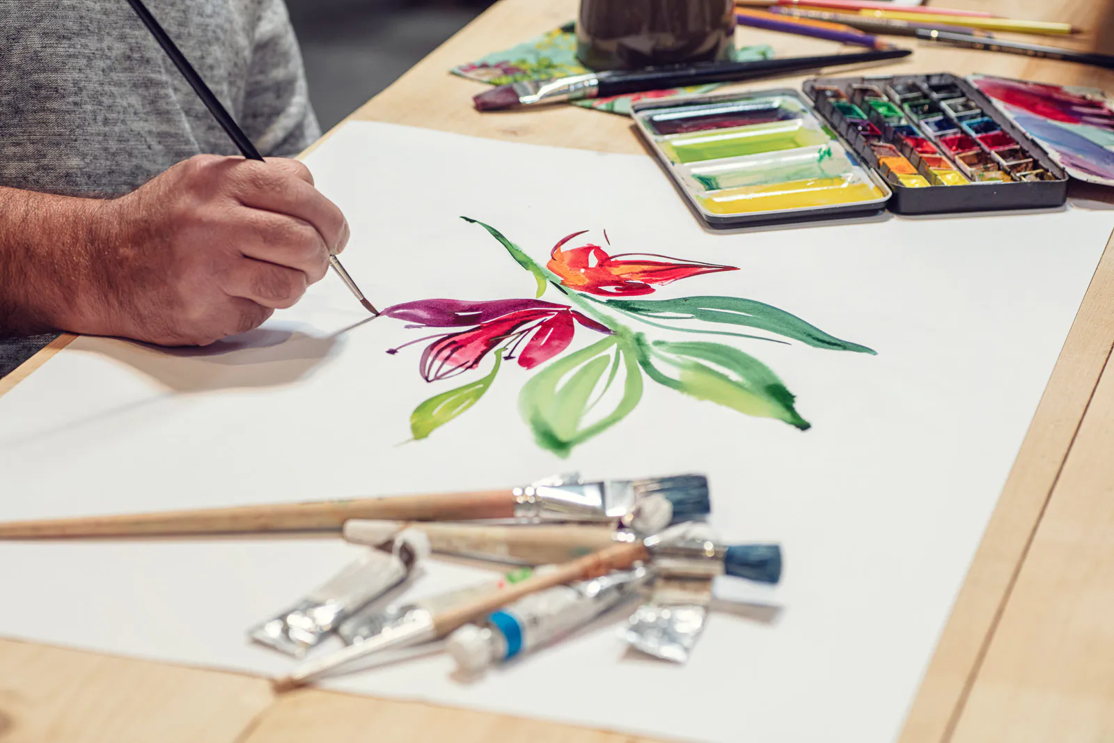 Person malt mit einem feinen Pinsel rote Blüten und grüne Blätter; Aquarellkasten und Pinsel liegen auf dem Holztisch.