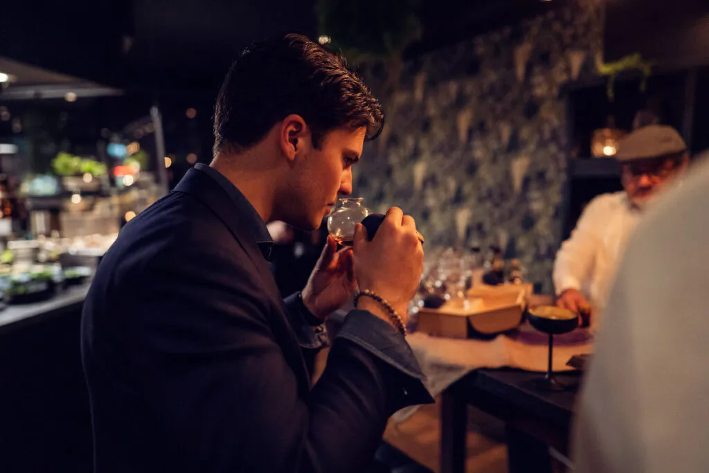 Ein Mann riecht an einem Glas mit bernsteinfarbenem Rum, im Hintergrund eine Bar in warmem, gedämpftem Licht.