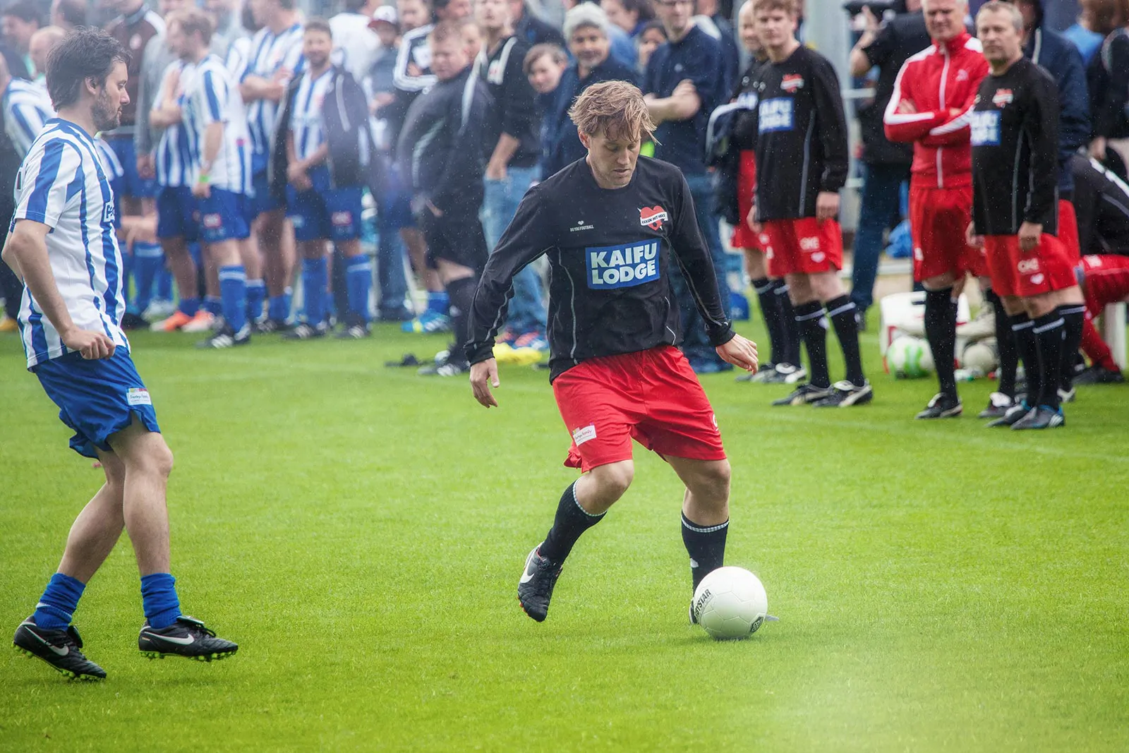 Fußballspieler in schwarzem Trikot und roter Hose führt den Ball auf dem Rasen, weitere Spieler stehen im Hintergrund.