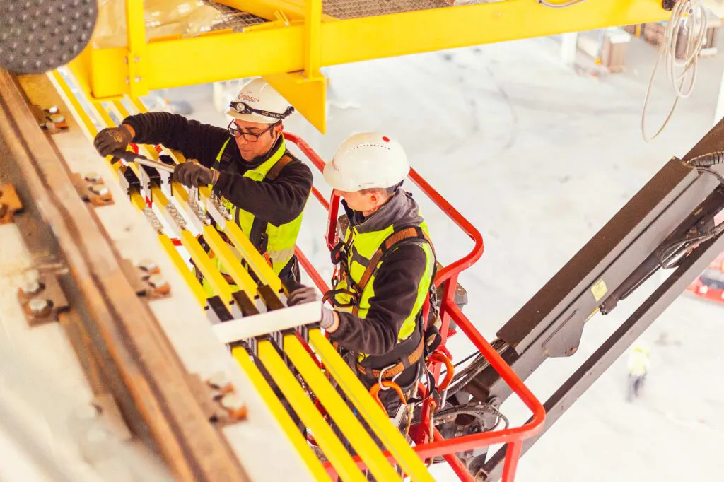 Zwei Arbeiter mit Helmen und Warnwesten montieren gelbe Stromschienen von einer Hebebühne in einer hellen Industriehalle.
