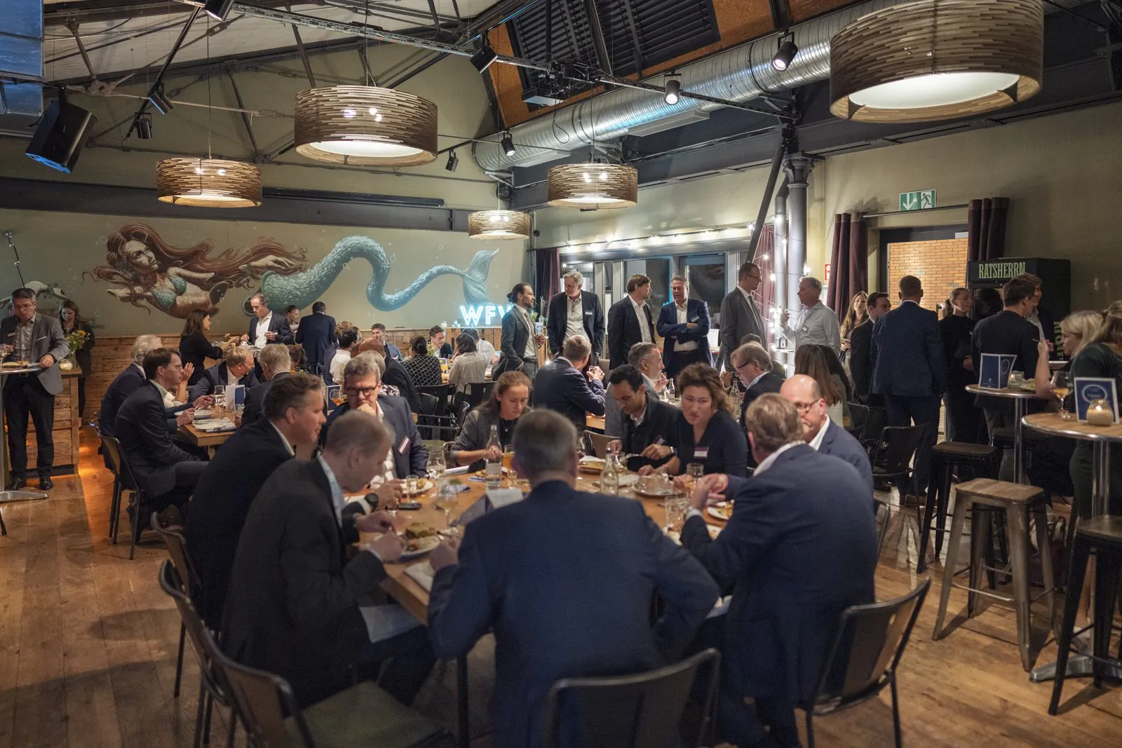 Gäste sitzen und stehen an Holztischen in einer warm beleuchteten Eventlocation mit Drachenmotiv an der Wand.