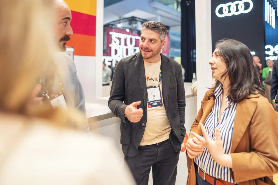 Mann im Awin-Shirt und dunklen Sakko im Gespräch mit einer Frau im braunen Blazer am hell beleuchteten Messestand.