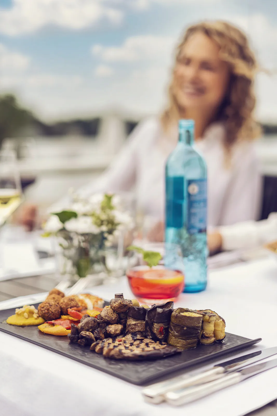 Grillgemüse und Getränke auf einem sonnigen Beachclub-Tisch, dahinter eine lächelnde Frau im weißen Hemd.