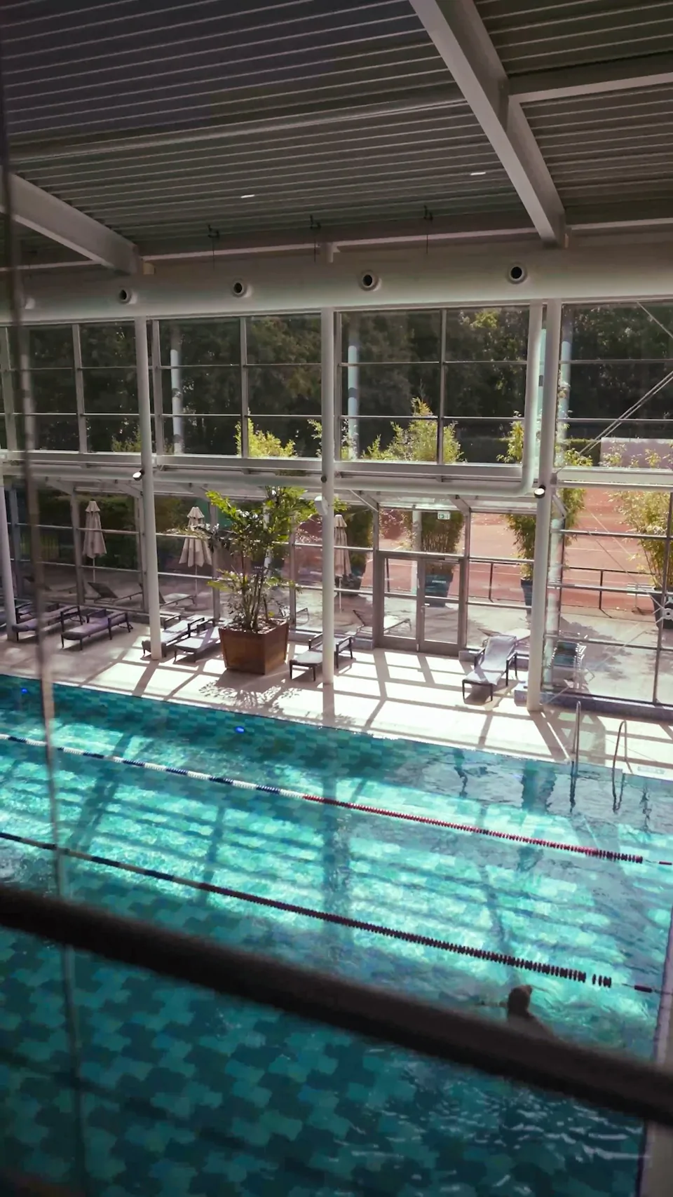 Sonnendurchflutetes Hallenbad mit türkisblauem Schwimmbecken, Schwimmbahnen, Liegen und großen Fensterfronten.