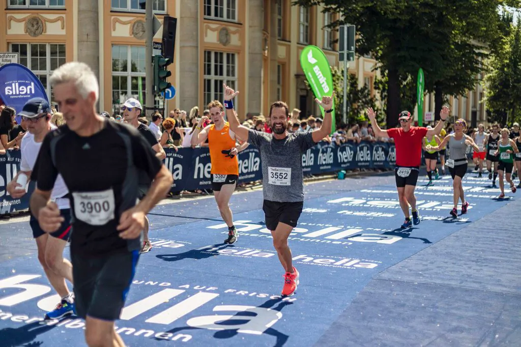 Läufer jubeln mit erhobenen Armen auf einer blauen Laufstrecke vor Zuschauern und Gebäuden im Sonnenlicht.