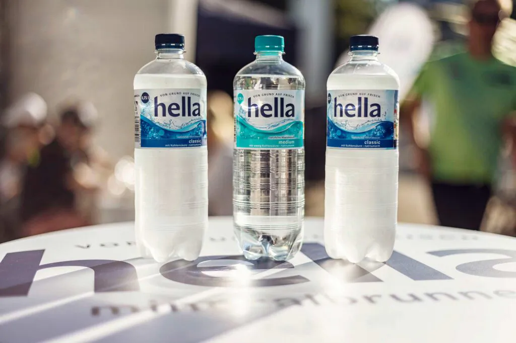 Drei hella-Wasserflaschen auf einem runden Tisch im Sonnenlicht, dahinter unscharfe Personen.