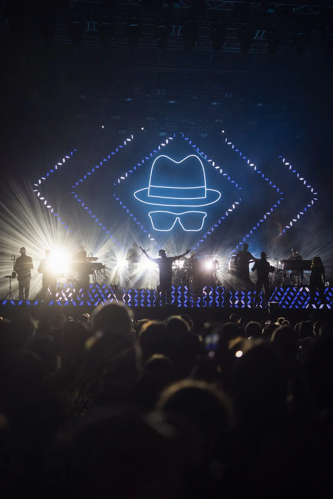 Band als Silhouetten im Gegenlicht auf der Bühne, dahinter leuchtende Hut- und Brillenkonturen, davor Publikum.